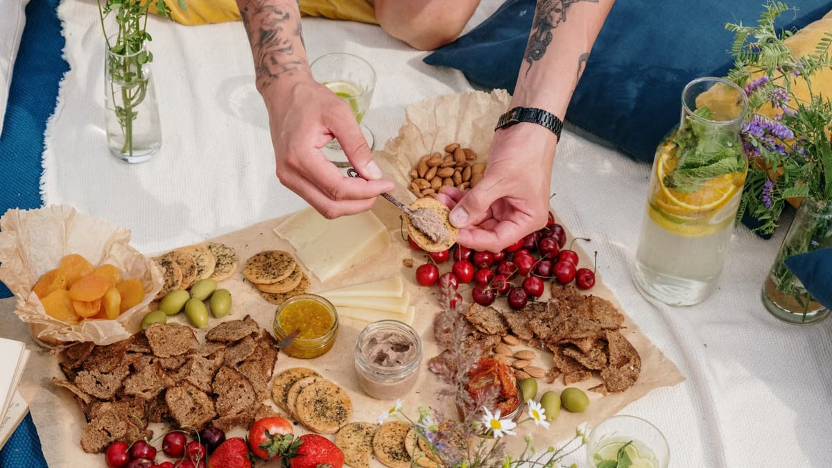 A colorful outdoor picnic featuring fruits, cheeses, and snacks on a blanket with plants in view.